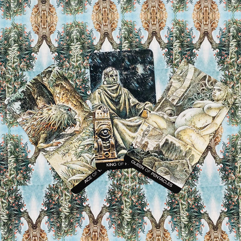 Tarot kortos iliustruotas rinkinys su Ace, King ir Queen of Pentacles kortomis ant smėlio‑mėtų ornamentuoto fono, artimas planas, centre trijų kortų kompozicija