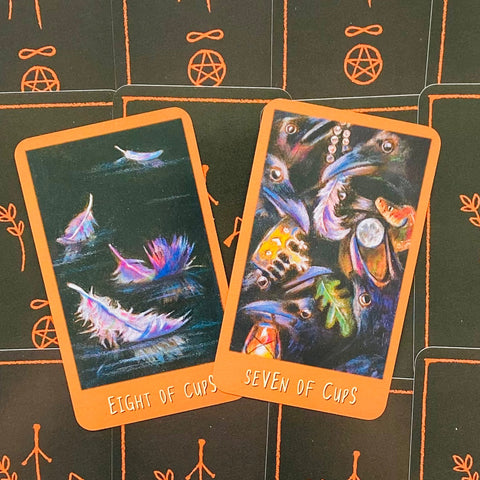 Tarot kortos su oranžiniu rėmeliu: „Eight of Cups“ ir „Seven of Cups“ padėtos ant juodo simbolinio fono, pasvirusios kampu