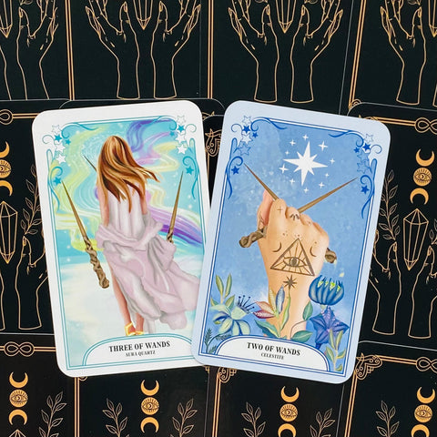 Taro kortos, dvi atverstos kortos (Three of Wands ir Two of Wands) su pastelinėmis iliustracijomis ir juodomis nugarėlėmis su auksiniais simboliais, artimas kampas