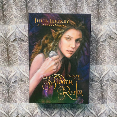 Tarot kortos "Tarot of the Hidden Realm" dėžutė su Julia Jeffrey ir Barbara Moore viršeliu, artimas moters portretas purpurinė–žalsva paletė