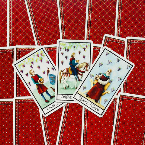Tarot kortos su raudonomis nugarėlėmis, centre trys atverstos Puodelių figūros — jaunuolis, riteris ir karalienė