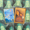 Tarot kortos — dvi demonstracinės kortos su mėlyna ir ruda kortelių rėmeliais bei auksine apdaila, parodytos iš viršaus ant žalsvo raštuoto fono