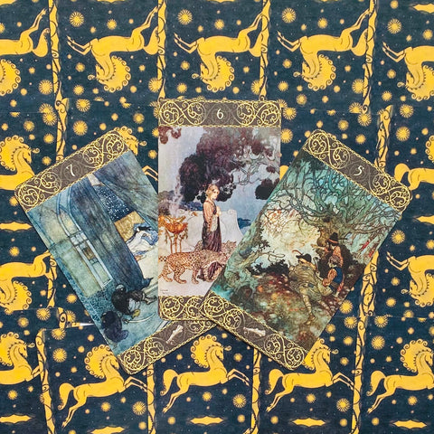 tarot kortos su vintage iliustracijomis, trijų kortų priekinės pusės iš arti ant tamsiai mėlyno audinio su geltonais žirgeliais, akcentuojant ornamentinį auksinį rėmą ir meninį stilių