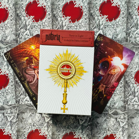 Tarot kortos Notoria: baltas dėklas su auksiniu saulės motyvu, dvi iliustruotos kortos ir raudonas viršelis fone