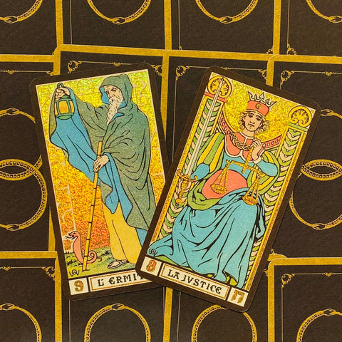 Tarot kortos Rider–Waite stiliaus su auksiniu blizgiu fonu, dvi kortos (Ermitas ir Teisingumas) padrikai padėtos iš viršaus