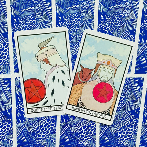 Tarot kortos Queen ir King of Pentacles kampu išdėstytos ant mėlynos raštuotos nugarėlės, matomas kortų dizainas ir spalvų derinys