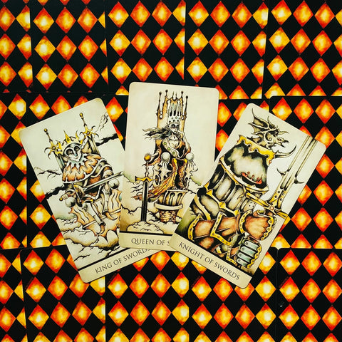 Tarot kortos su oranžinėmis rombų nugarėlėmis — trys atvertos karališkos kortos (Karalius, Karalienė, Riteris) fanuotos, rodant piešinių ir spalvų detales