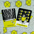 Tarot kortos su geltonai‑juodais iliustruotais dizainais, matomi Ten of Ray Guns ir Nine of Ray Guns priekiai bei žvaigždėtas nugarėlių raštas, demonstruoja kortų stilių pirkėjui