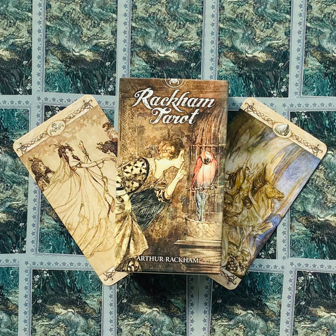 Rackham Tarot kortos: dėžutė su Arthur Rackham viršeliu ir dvi pasvirusios kortos ant žalsvai mėlyno iliustruoto fono