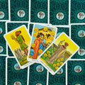 Tarot kortos Rider‑Waite stiliaus: trys atverstos pentaklių kortos (Page, Ten, Nine) išdėstytos fanu ant žalsvai mėlyno ornamentuoto nugarėlių fono