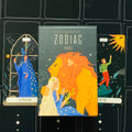 Tarot kortos Zodiac – dėžutės priekinis viršelis tamsiai žalias su oranžiniu liūtu, šalia The Magician ir The Fool kortos