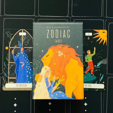 Tarot kortos Zodiac – dėžutės priekinis viršelis tamsiai žalias su oranžiniu liūtu, šalia The Magician ir The Fool kortos