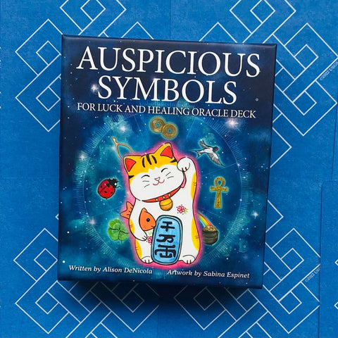 Orakulo kortų rinkinys su mėlyna dėže ir besišypsančiu maneki‑neko talismanu bei įvairiais sėkmės simboliais ant viršelio