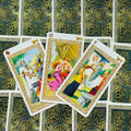 Tarot kortos su keltų nugarėlėmis, trys atvertos iliustruotos kortos fane, artimas planas akcentuoja auksinį rėmelį ir ryškias spalvas