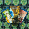 tarot kortos Mystic Spiral dėžutė su mėlynais ir auksiniais viršelio piešiniais, šalia dvi atverstos kortos ir žalio-juodo ornamentuoto fonas