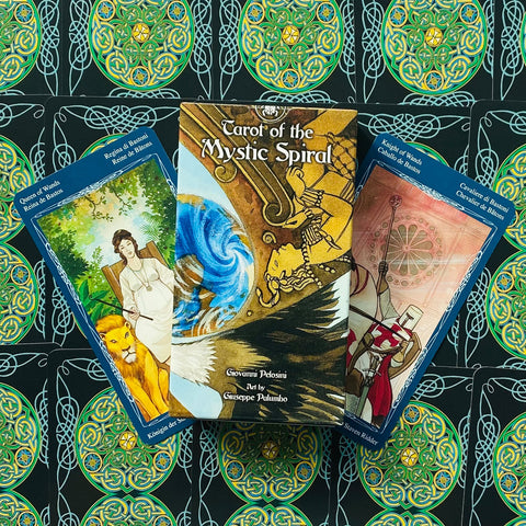 tarot kortos Mystic Spiral dėžutė su mėlynais ir auksiniais viršelio piešiniais, šalia dvi atverstos kortos ir žalio-juodo ornamentuoto fonas