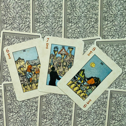 Tarot kortos Rider‑Waite stiliaus, kremiškomis kraštinėmis, trijų taurų kortų išklotinė ant rašto nugarėlių, arti fotografuota