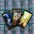 Tarot kortos giliai mėlynais ornamentuotais nugarėmis; trys atverstos juodo‑aukso rėmelių kortos su gotikinėmis ir šviesiomis scenomis, parodytos ventiliatoriumi