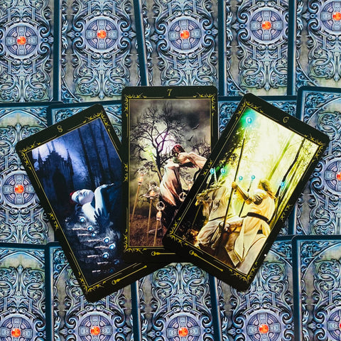 Tarot kortos giliai mėlynais ornamentuotais nugarėmis; trys atverstos juodo‑aukso rėmelių kortos su gotikinėmis ir šviesiomis scenomis, parodytos ventiliatoriumi