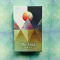 The Field Tarot kortų rinkinys dėžutėje, frontinis vaizdas su geometriniais trikampiais, saulės/menės simboliu ir žaliu‑melsvu fonu