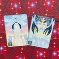 Tarot kortos su pastelinių atspalvių angeliškomis iliustracijomis, dvi atviros kortos parodytos ant raudono ornamentuoto fono