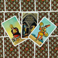 Tarot kortos Rider–Waite stiliaus, trys atviros pentaklių kortos iš viršaus ant ornamentuotos raudono‑juodo nugarėlės, ryškūs spalvoti motyvai