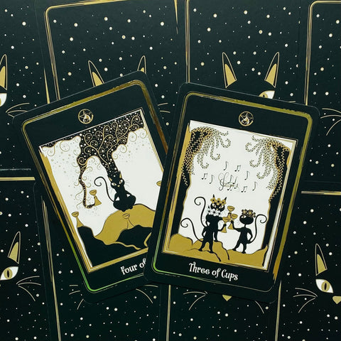Tarot kortos katėmis, juodos ir auksinės, dvi išverstinės kortos (Three of Cups ir Four of Cups) pateiktos ant žvaigždėto fono