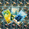 Tarot kortos meninės iliustruotos, pilkai apvadais, trys atvertos kortos centre ant tamsios paukštinės nugarėlės dekoracijos