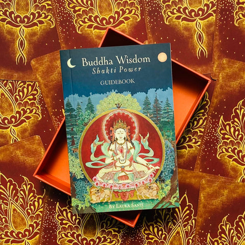 Knyga Buddha Wisdom: Shakti Power — mėlynas viršelis, aut. Laura Santi, dedama į atvirą oranžinę dovanų dėžutę su raudonų kortų nugarėlių raštais