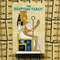 Egipto tarot kortų rinkinys su smėlio spalvos papiruso dėklu, priekyje sėdinčios faraonės iliustracija.