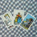 tarot kortos Rider–Waite komplekte — trys atverstos kortos (Ace of Swords, King of Cups, Queen of Cups) ant mėlyno languoto fono, plokščias išplanavimas