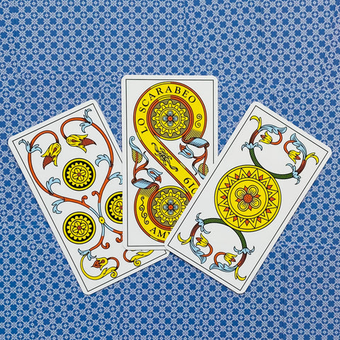 Tarot kortos su monetų rūšies raštais, trys ornamentuotos geltonos monetos išdėstytos ant mėlyno raštuoto fono