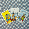 tarot kortos Rider‑Waite stiliaus, trys kardų kortos (VII, VI, V) ant mėlyno‑baltos languotos staltiesės, artimas plokščias išdėstymas