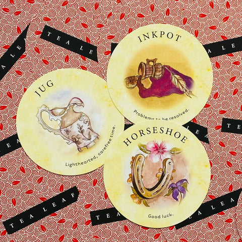 Orakulų kortos Tea Leaf — trys apvalios gelsvos kortos (Jug, Inkpot, Horseshoe) išdėstytos ant raudono raštuoto fono, pavyzdinis simbolių variantas
