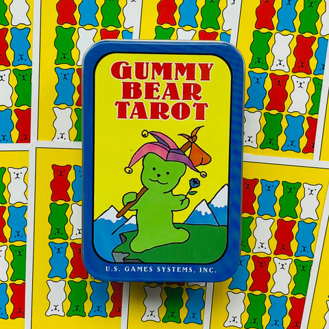 Gummy Bear Tarot kortų rinkinys metalinėje mėlyno rėmo dėžutėje su geltonu viršeliu ir žaliu meškiuku, fone spalvotos kortų nugarėlės