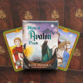 Mists of Avalon orakulo kortų komplektas, pakuotė priekyje ir dvi atvertos iliustruotos kortos šonuose