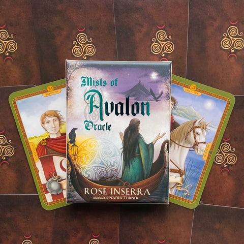 Mists of Avalon orakulo kortų komplektas, pakuotė priekyje ir dvi atvertos iliustruotos kortos šonuose