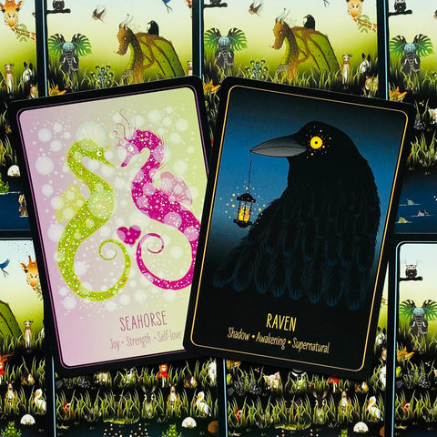 Orakulų kortos, dvi atverstos kortos: rožinė–žalia Seahorse ir juoda Raven su žibintu, parodytos ant iliustruoto, pasakiško fono, kad pademonstruotų dizaino variantus