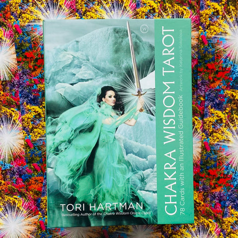 Chakra Wisdom Tarot kortų dėžutė su žalsvu viršeliu, moters figūra laikanti kardą, padėta ant spalvingo audinio fono