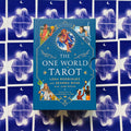 Tarot kortos The One World Tarot mėlyname dėkle su autoriaus vardais ir iliustracijomis, fotografuotos ant mėlyno žvaigždėto kortų fono