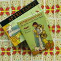 After Tarot kortų rinkinys su žalia „Afterthoughts“ vadovu ir juoda dėžute, pateiktas ant dekoratyvių kortų nugarėlių fono