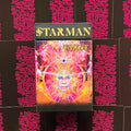 Starman tarot rinkinys su spalvingu psichodeliniu viršeliu ir juodu dangteliu auksiniais užrašais, matomas iš priekio ant rožinių kortų nugarėlių fono