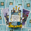 Weiser Tarot kortų dėžutė centre ant išskleistų kortų fono; ryškūs mėlyni ir geltoni tonai, trejų taurų motyvas