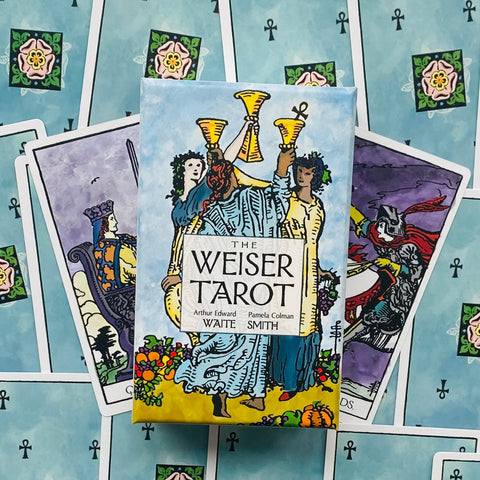 Weiser Tarot kortų dėžutė centre ant išskleistų kortų fono; ryškūs mėlyni ir geltoni tonai, trejų taurų motyvas