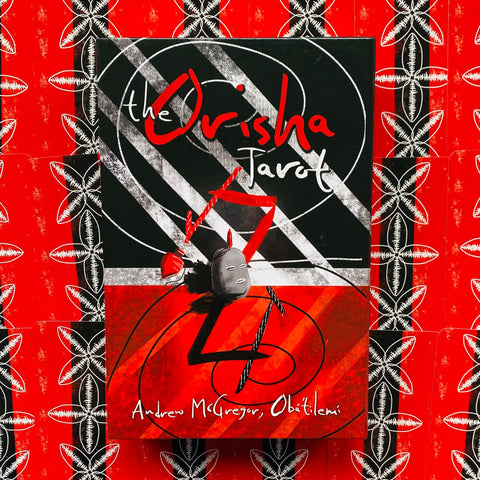 Orisha tarot dėžutė su juoda–raudonu viršeliu ir autoriaus parašu, parodyta priekinė pusė ant raudono raštuoto fono