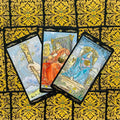 Tarot kortos su juodai auksinėmis ornamentuotomis nugarėlėmis: trys atvertos kortos (Lazdos, Karalius, Karalienė Pentaklių) elegantiškai išskleistos virš raštuoto fono