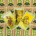 Tarot kortos Wild Unknown pentacles rinkinys: trys geltonos monetų kortos ant žaliai‑raudono ornamentuoto audinio, išdėstytos iš viršaus