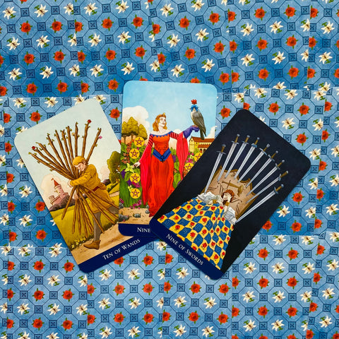 Tarot kortos komplektas su trimis atverstomis kortomis (Dešimt lazdų, Devynios pentaklės, Devynios kardai) ant mėlyno gėlių fono, pasvirusiu išdėstymu