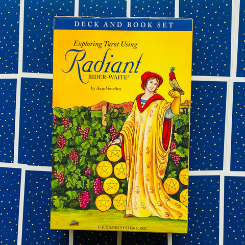Radiant Rider‑Waite taro rinkinys geltonoje dėžutėje su knyga, priekyje figūra ir pentaklių monetos, fone mėlyni kortų nugarėlių raštai