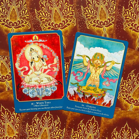 orakulo kortos: dvi atverstos kortos (White Tara ir Garuda) ant raudono-aukso ornamentuoto fono, plokščias iš viršaus